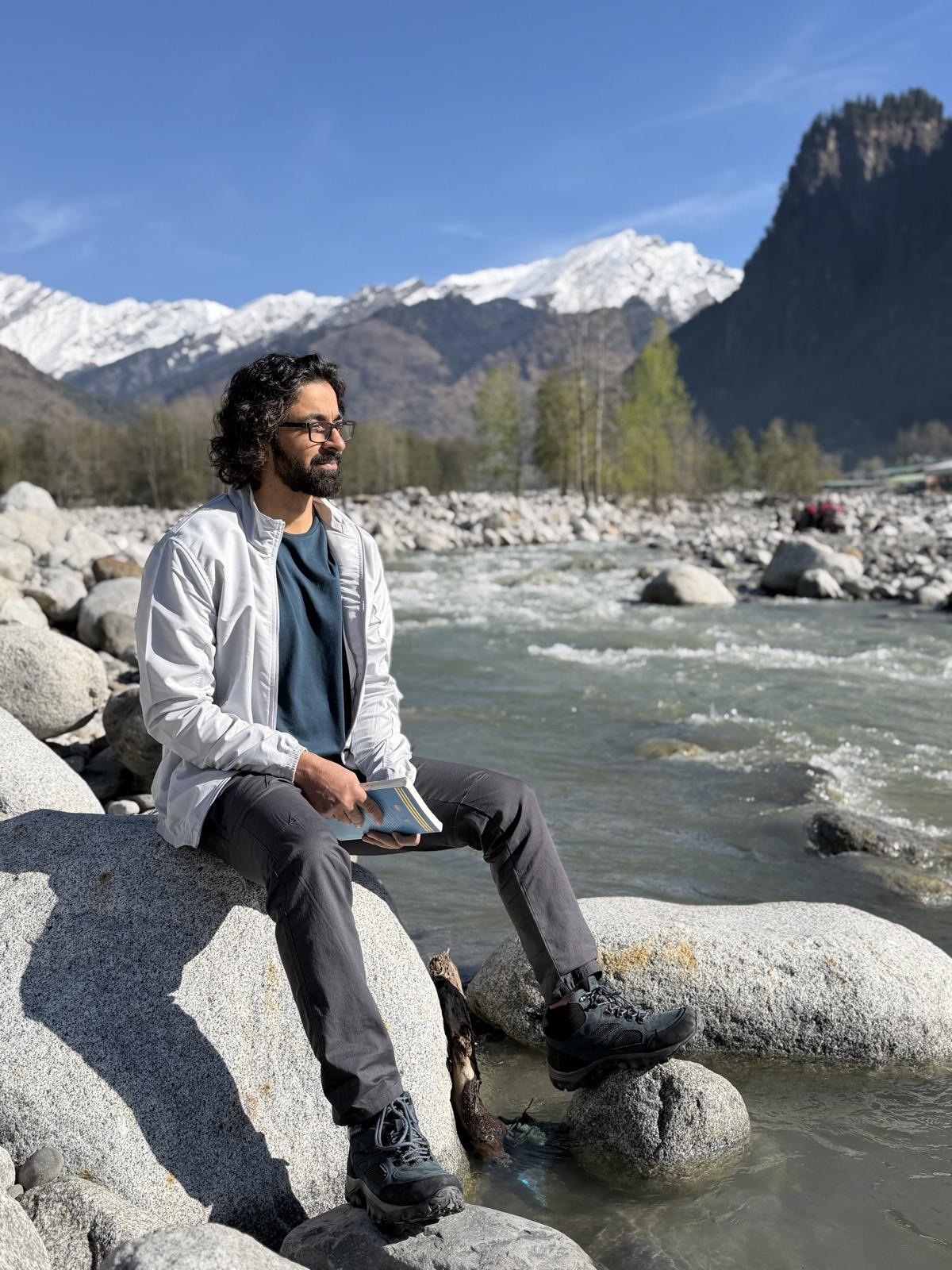 Deepak Atri sitting by a mountain river with a book